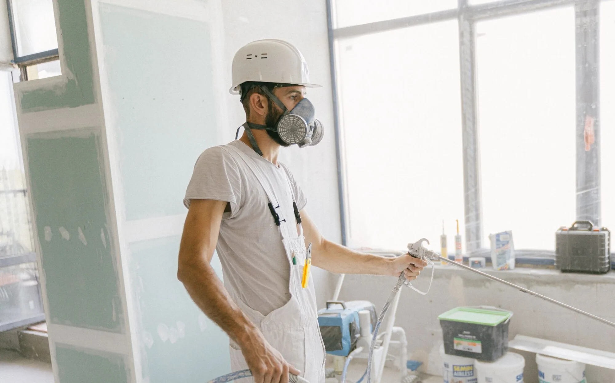 Professional painter in white overalls, hard hat, and respirator running an airless spray rig inside a bright renovation site