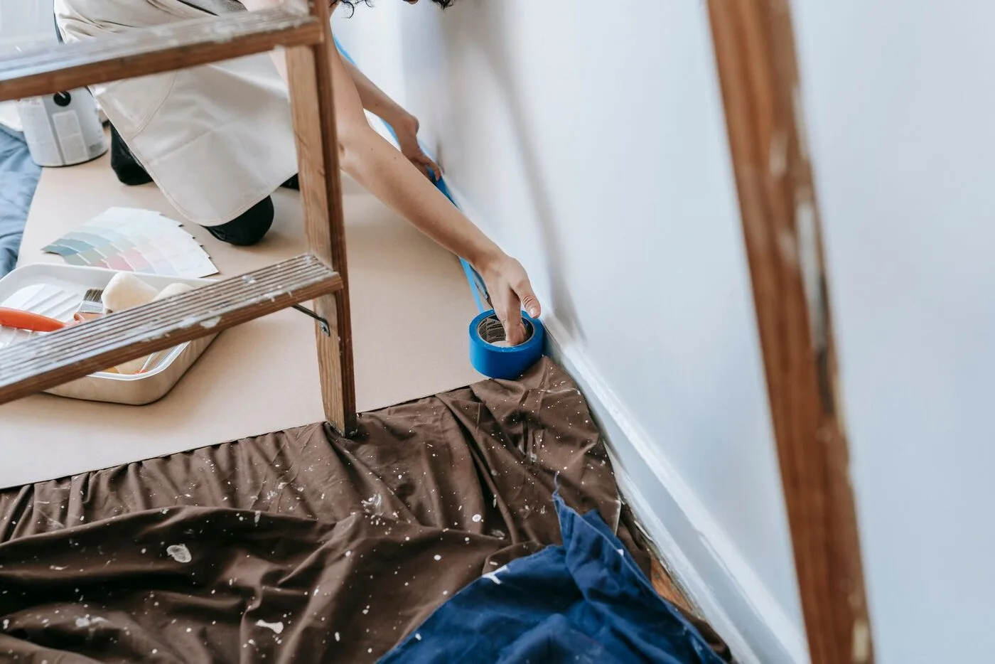 Painter masking baseboards with tape over protected floors before painting begins